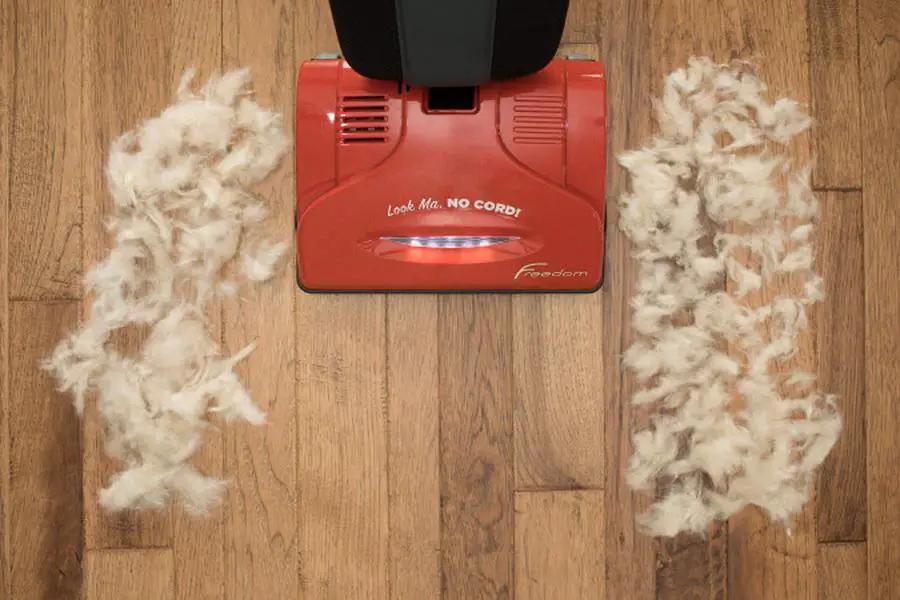Simplicity S10CV Freedom red cordless upright vacuum on wood floor with dust clumps on either side.