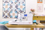 Bernette B59 sewing and embroidery machine in white on a wooden table with a quilt backdrop, by BERNINA.