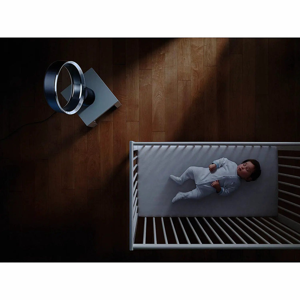 Dyson AM06 10in desk fan in white on a wooden floor beside a sleeping baby in a crib.