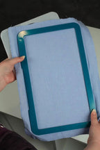 Embroidery hoop with teal frame holding light blue fabric, shown being held by two hands
