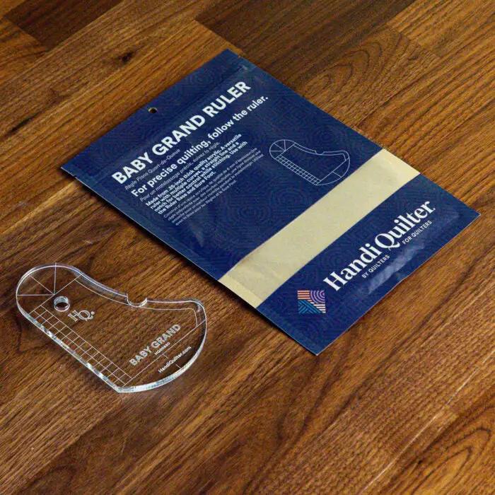 Baby Grand Quilting Ruler by HandiQuilter, transparent acrylic quilting ruler beside blue instructional booklet.