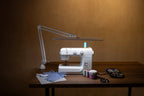 Daylight Lumi LED Task Lamp with a gray metal adjustable arm on sewing table near thread spools.