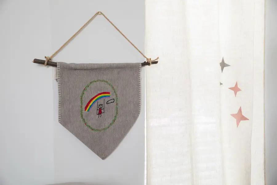 Embroidery banner in grey fabric with a rainbow motif, hung on a wooden rod with twine.