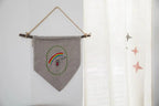 Embroidery banner in grey fabric with a rainbow motif, hung on a wooden rod with twine.