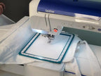 Embroidery machine with blue top, fabric in a square hoop; Brother brand visible