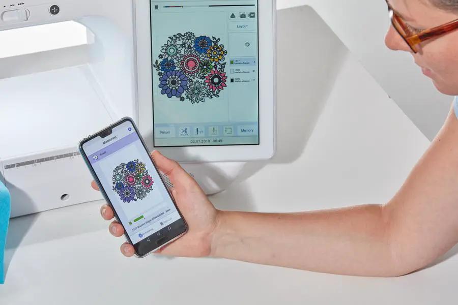 Brother Stellaire Innov-is XJ1 sewing and embroidery machine, white, with smartphone displaying embroidery design