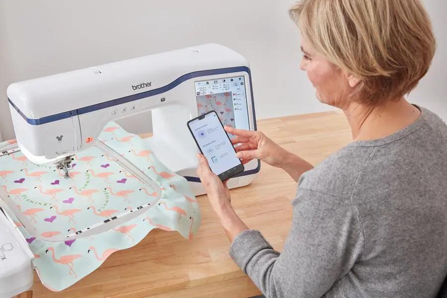 Brother Stellaire Innov-Is XE1 embroidery machine, white with blue trim, on a table with flamingo fabric and smartphone control.