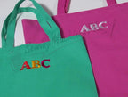 Two tote bags in bright green and pink with embroidered ABC logos