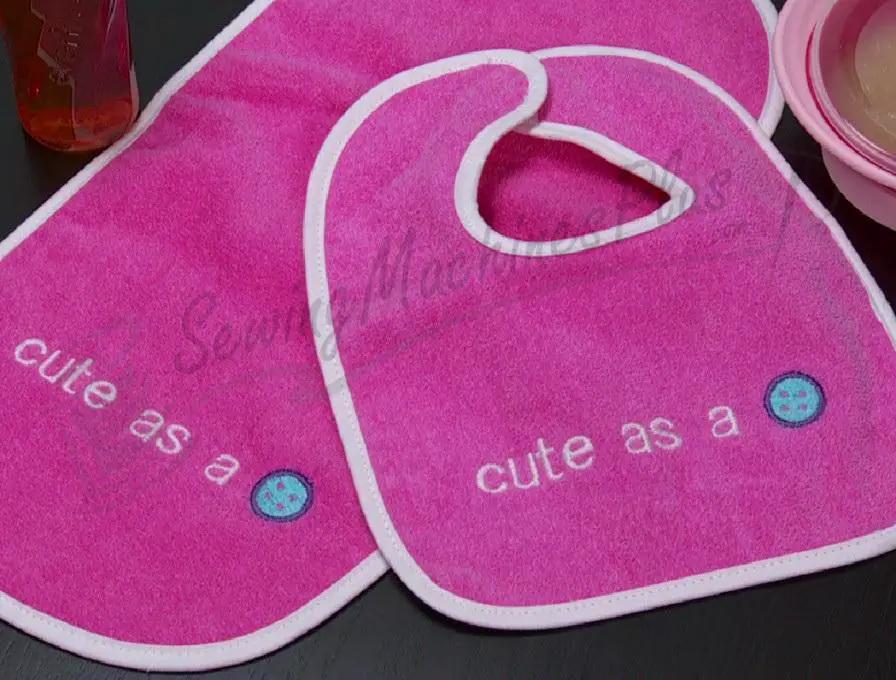 Pink baby bibs with white trim, featuring the phrase cute as a and blue button graphics.