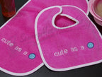 Pink baby bibs with white trim, featuring the phrase cute as a and blue button graphics.